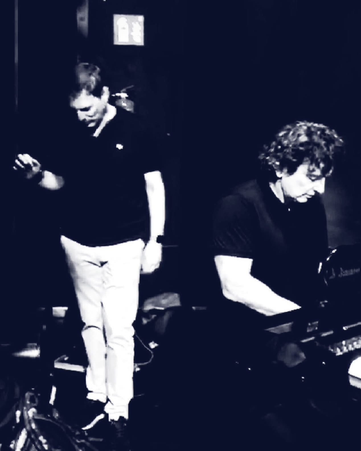 Sergio Cano Ortiz de pie y Jim Papoulis al piano, en blanco y negro.