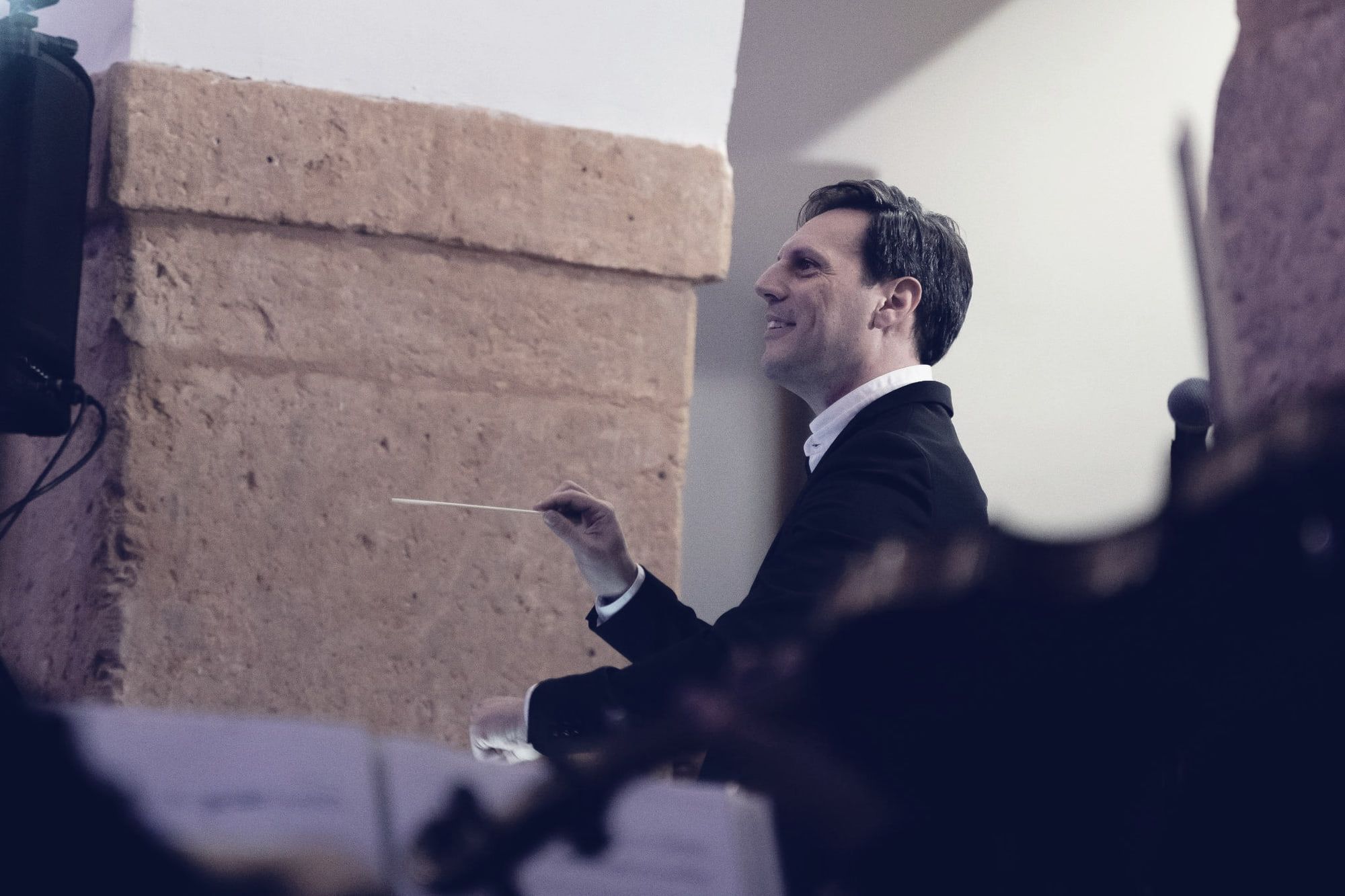 Sergio Cano Ortiz dirigiendo a la orquesta con la batuta, sonriendo, junto a una columna de piedra.